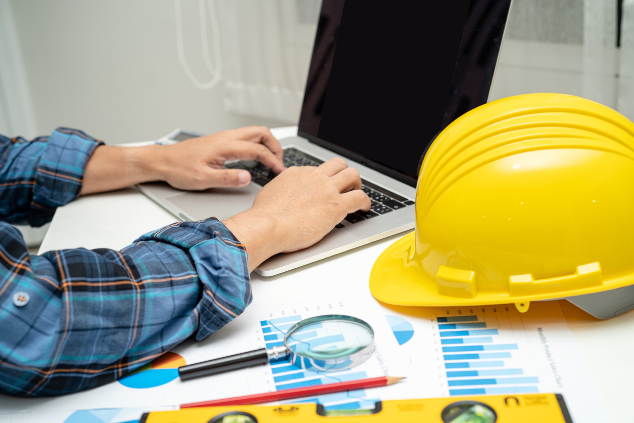 Hands on laptop with blueprints and yellow hard hat nearby